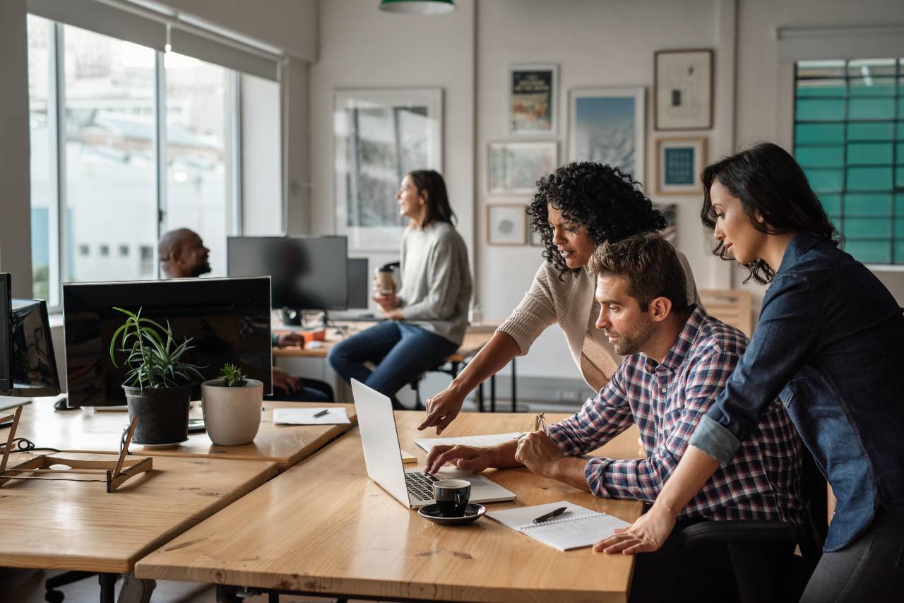 Shows a team of technology company workers, a man and two women, crowded aroud a laptop computer at a desk in a modern looking office.